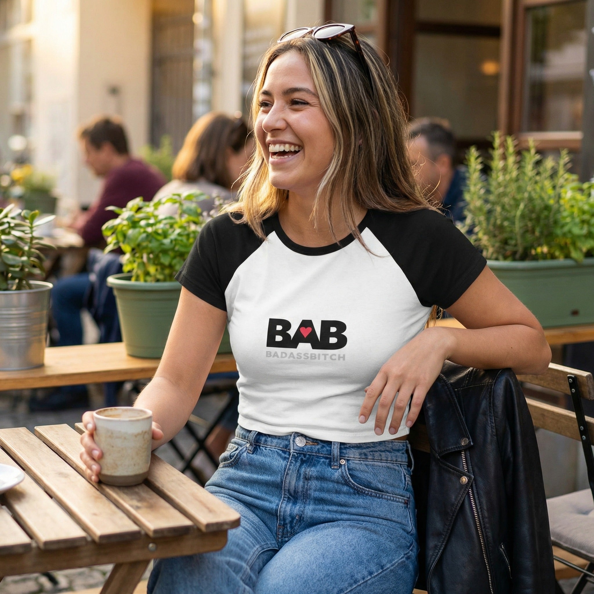 Woman sitting at an outdoor cafe table holding a mug, wearing a t-shirt with 'B.A.B. BADASS BITCH' text.