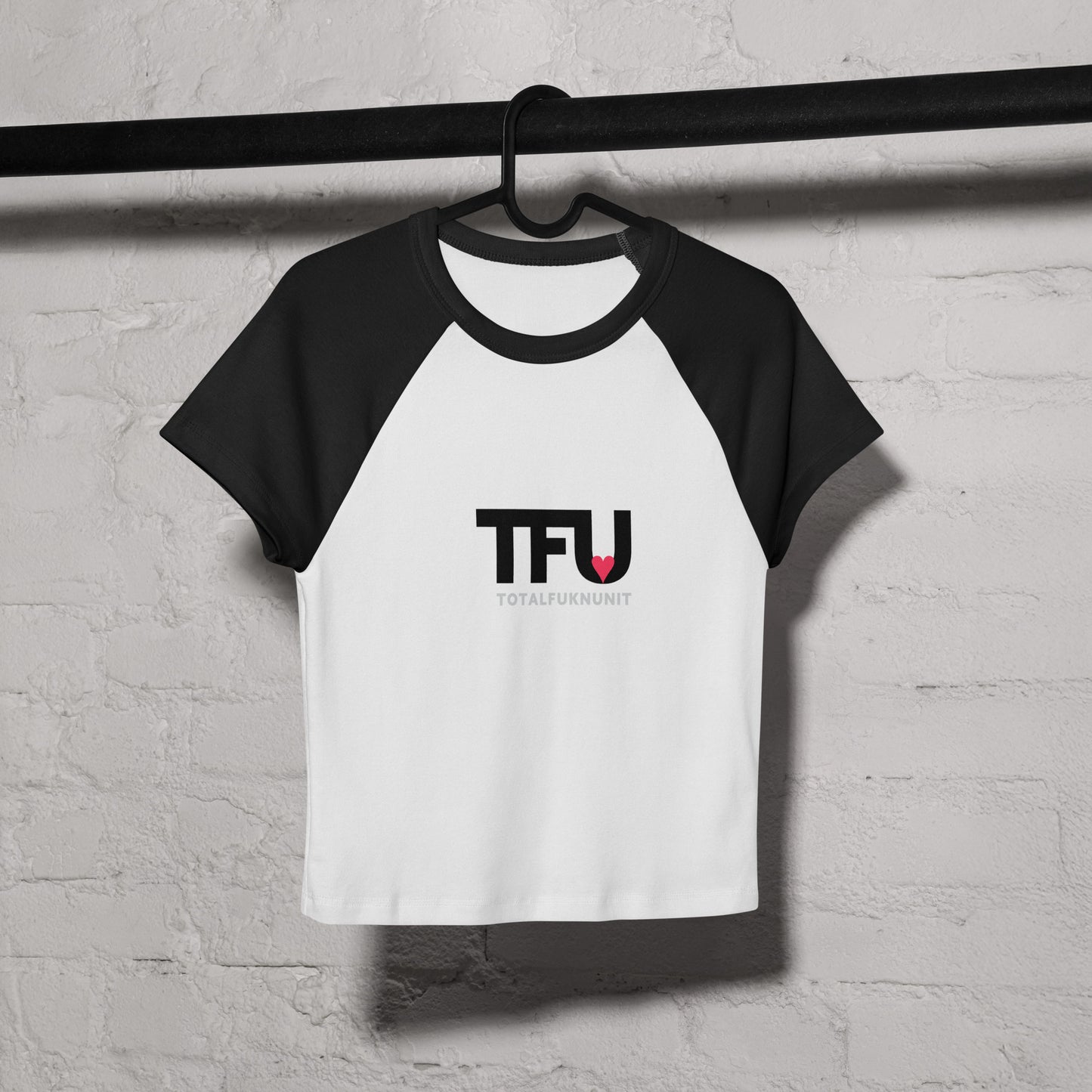 Black and white baby tee shirt with 'TFU' logo on a hanger against a grey brick wall.