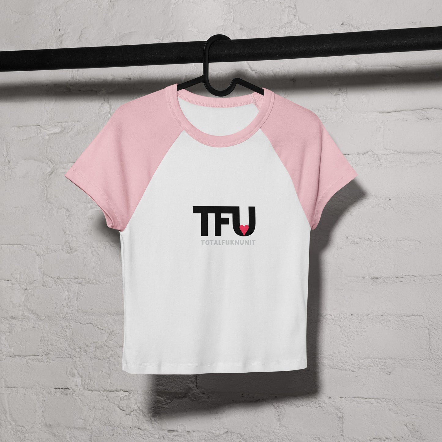 Pink and white baseball shirt with 'TFU' logo on a hanger against a gray brick wall.